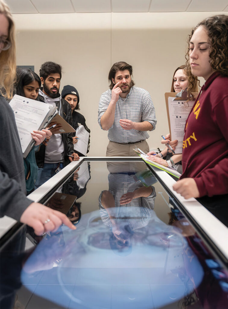 A professor lecturing with a group of students surrounding the Anatomage Table.