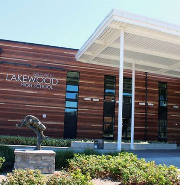 Main entrance of Lakewood High School with modern wood-panel architecture, large windows, metal dolphin statue, and landscaped bushes under a clear blue sky.